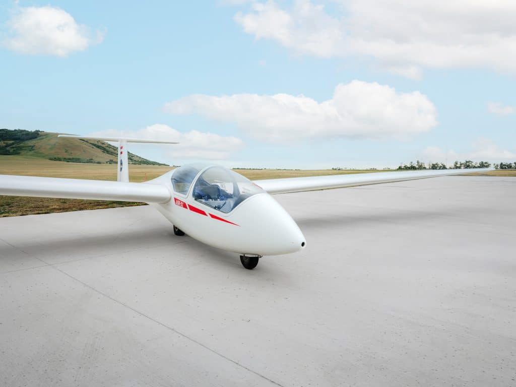 Segelflugzeug in Weiß mit rotem Streifen auf dem Vorfeld am Flugplatz Spitzerberg, Seitenansicht mit geschlossener Glaskabine, dahinter weite Wiesenlandschaft und Hügel unter leicht bewölktem Himmel