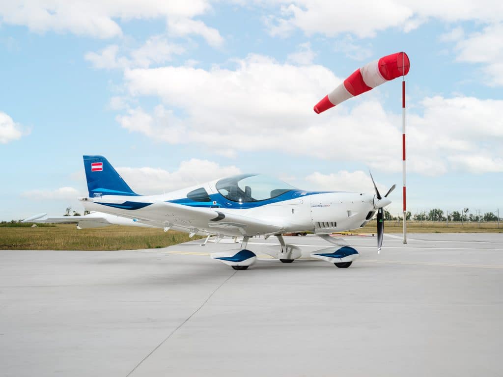 Modernes Kleinflugzeug (Bristell B23) in Blau-Weiß auf dem Vorfeld am Flugplatz, seitliche Ansicht mit Propeller und Kabine, daneben ein rot-weiß gestreifter Windsack unter leicht bewölktem Himmel
