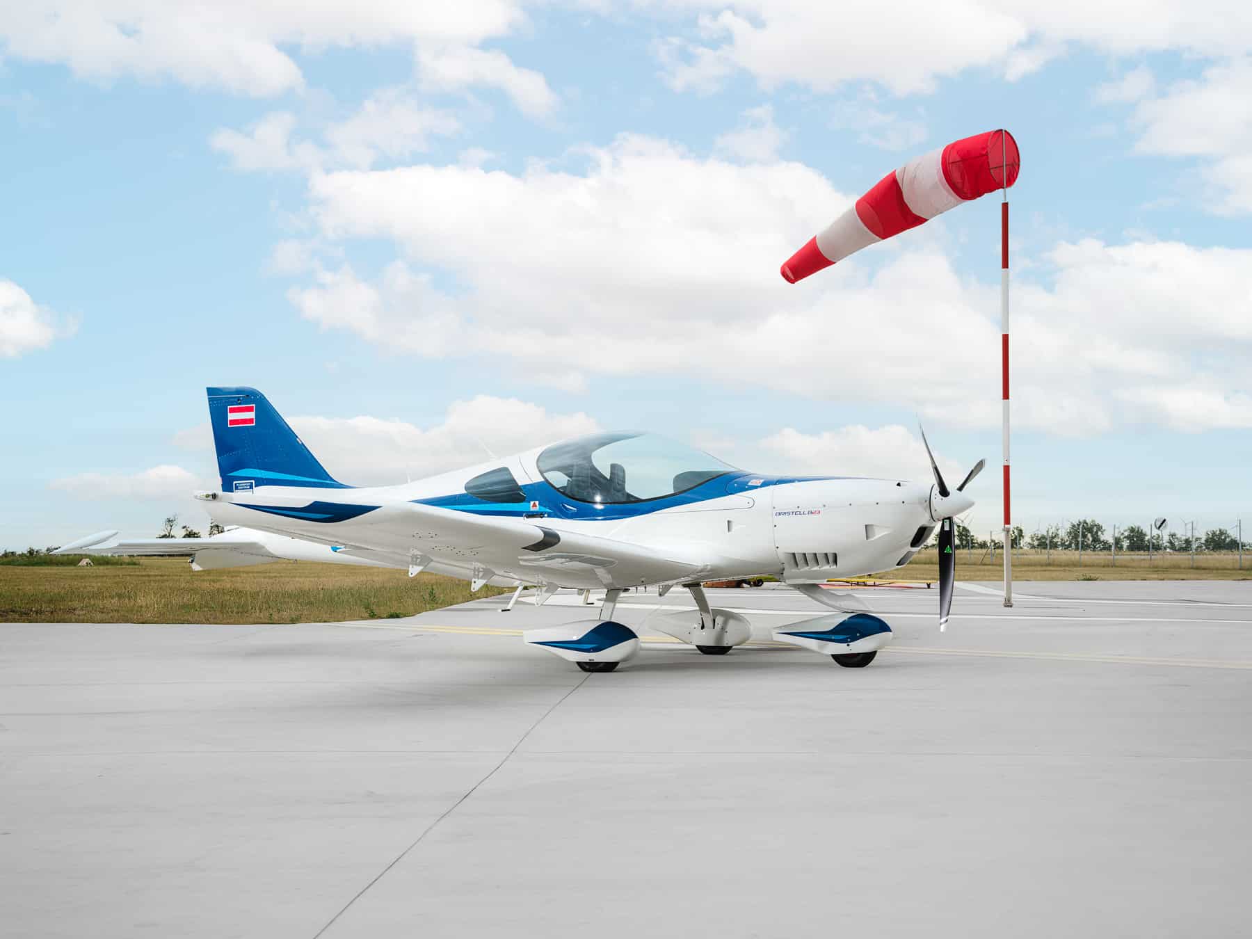 Modernes Kleinflugzeug (Bristell B23) in Blau-Weiß auf dem Vorfeld am Flugplatz, seitliche Ansicht mit Propeller und Kabine, daneben ein rot-weiß gestreifter Windsack unter leicht bewölktem Himmel