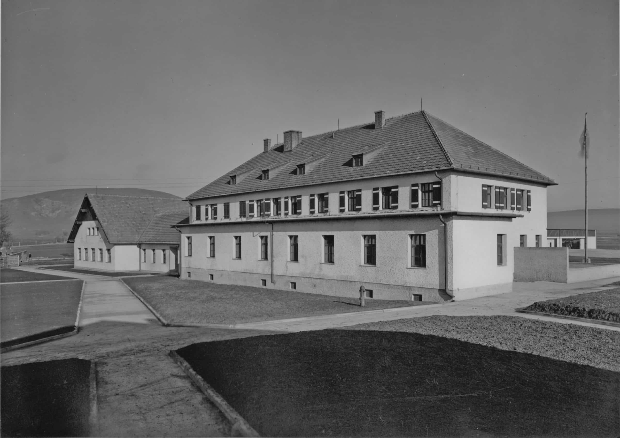 Historische Schwarz-Weiß-Aufnahme eines Gebäudes am Spitzerberg mit langgestrecktem Hauptbau, Satteldach und gleichmäßig angeordneten Fenstern, daneben Nebengebäude und offene Landschaft im Hintergrund