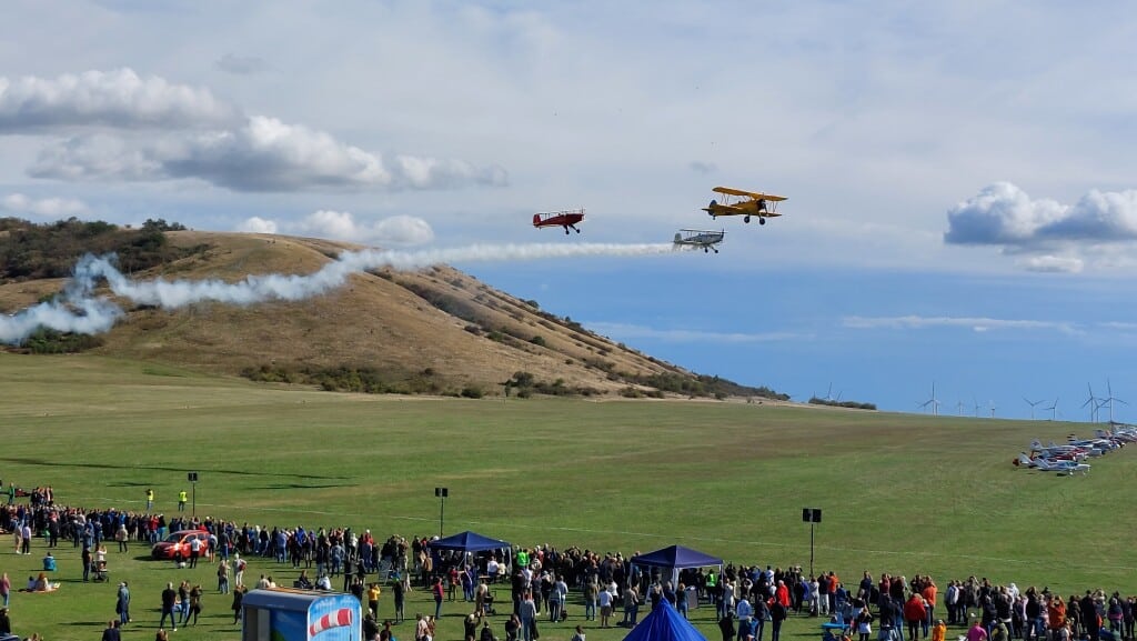 Mehrere historische Doppeldecker-Flugzeuge fliegen in Formation über dem Flugplatz Spitzerberg, eines zieht eine Rauchspur, darunter zahlreiche Zuschauer auf der Wiese sowie abgestellte Flugzeuge am Rand der Fläche