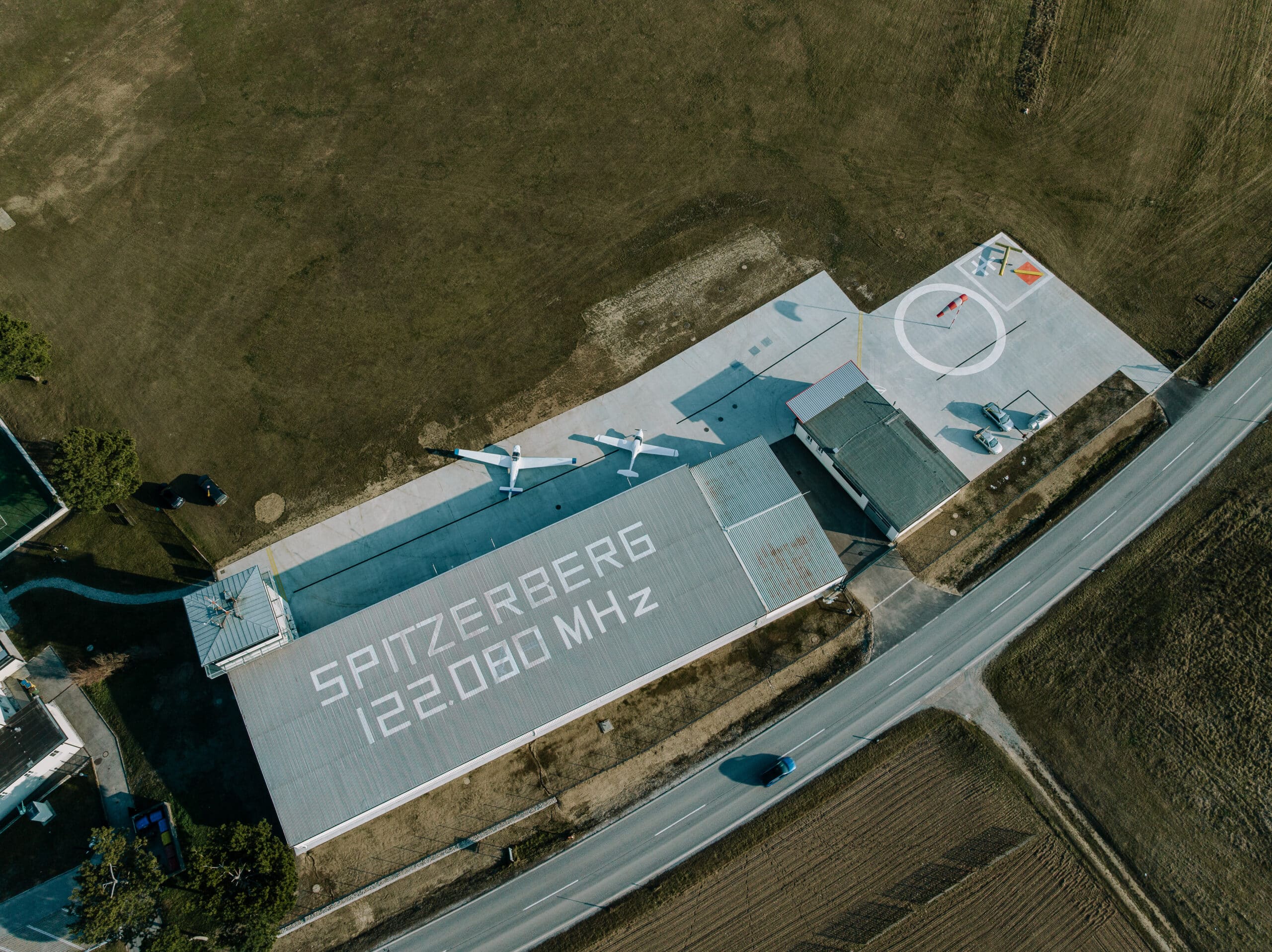 Luftaufnahme des Flugplatzes Spitzerberg mit Hangar (Aufschrift „Spitzerberg 122.080 MHz“), abgestellten Flugzeugen, Tower, Windsack und angrenzender Straße im ländlichen Gebiet