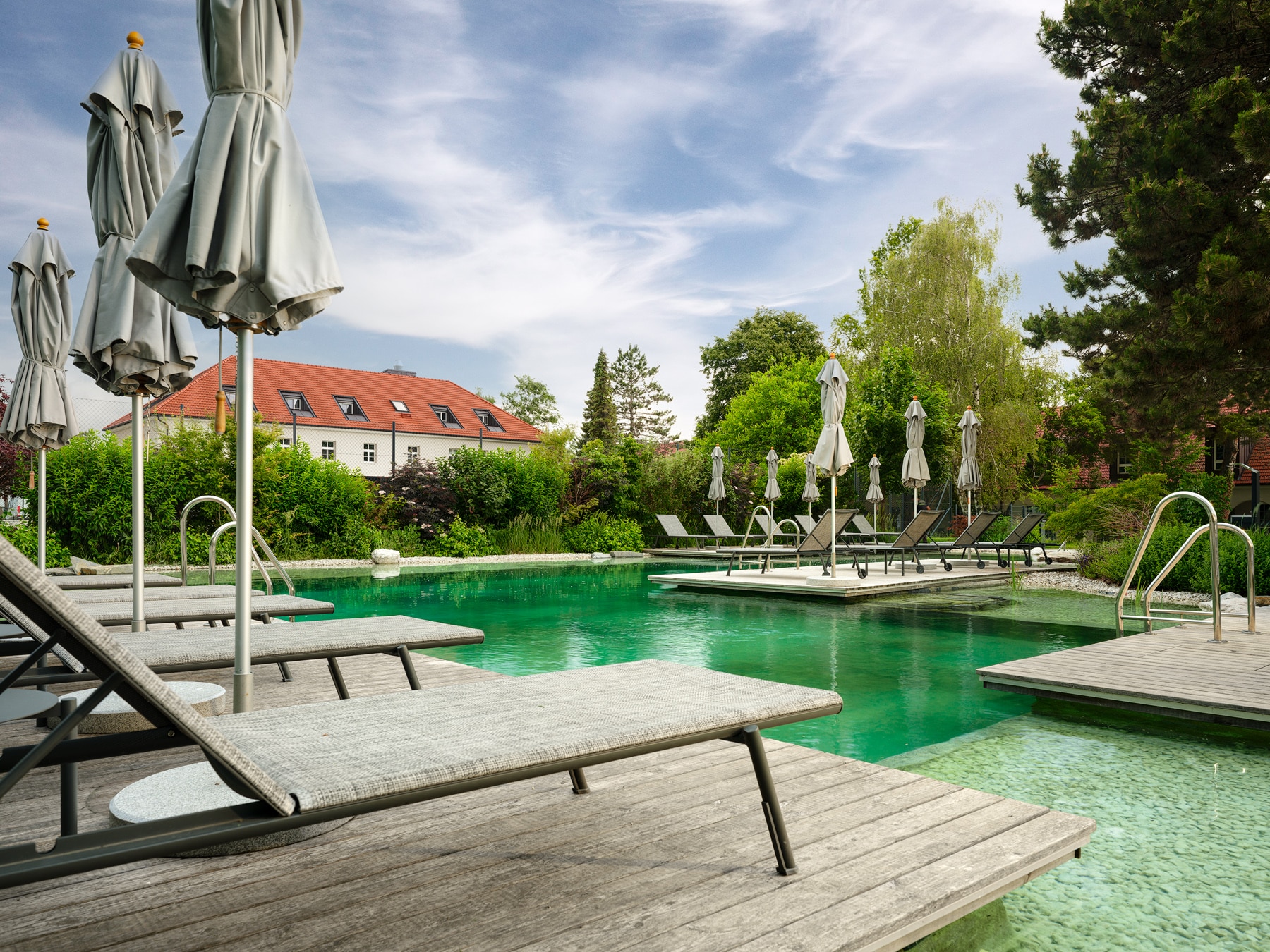 Naturpool mit Holzdeck, Liegen und geschlossenen Sonnenschirmen, umgeben von Gartenanlage und Gebäuden in ruhiger Umgebung am Spitzerberg