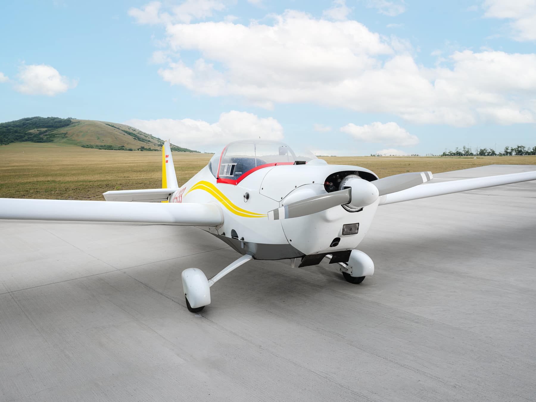 Weißes Kleinflugzeug mit gelben Streifen auf dem Vorfeld am Flugplatz Spitzerberg, seitliche Frontansicht mit Propeller, dahinter offene Wiesenlandschaft und Hügel unter bewölktem Himmel